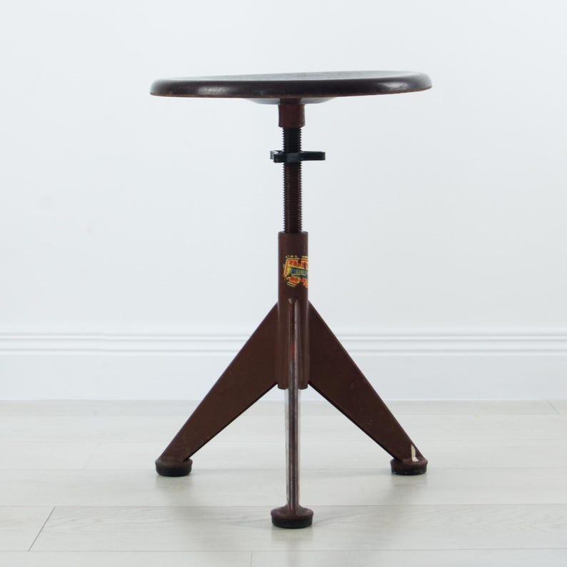 K&ouml;nnte beinhalten: Ein brauner Industriehocker mit rundem Sitz und dreibeinigem Untergestell. Der Hocker verf&uuml;gt &uuml;ber eine zentrale, mit Gewinde versehene Metalls&auml;ule zur H&ouml;henverstellung mit einem schwarzen Kunststoffknopf. Ein kleines Etikett ist sichtbar.