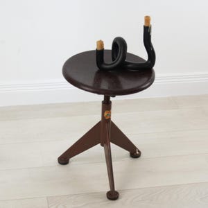K&ouml;nnte beinhalten: Ein dunkelbrauner, dreibeiniger Hocker mit runder Sitzfl&auml;che und verstellbarem Mittelpfosten. Auf der Sitzfl&auml;che steht ein schwarzer, geschwungener, r&ouml;hrenf&ouml;rmiger Kerzenhalter mit zwei hellen, teilweise abgebrannten Wachskerzen.