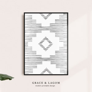 May include: Black and white geometric art print in a black frame. The design features horizontal and diamond-shaped patterns. The text "GRACE & LAGOM modern printable design" is at the bottom of the print. The artwork is displayed against a light pink wall.