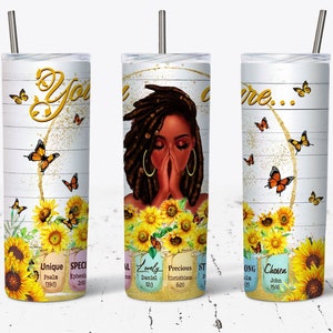 May include: A white tumbler with a black woman with dreadlocks praying, surrounded by sunflowers and butterflies. The tumbler has the words "You are..." and bible verses on it.