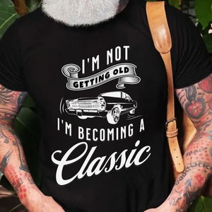 Könnte beinhalten: Schwarzes T-Shirt mit dem weißen Text "I'M NOT GETTING OLD, I'M BECOMING A CLASSIC". Unter dem Text befindet sich eine Grafik eines klassischen Autos. Das T-Shirt hat einen Rundhalsausschnitt und kurze Ärmel.