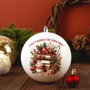 Op de afbeelding: Witte kerstbal met de tekst "FULLY BOOKED FOR CHRISTMAS". De bal heeft een stapel boeken, hulst, een zuurstok en een kopje warme chocolademelk. Een kleine rode bal staat op de voorgrond.
