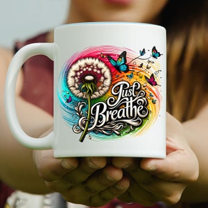 K&ouml;nnte beinhalten: Wei&szlig;e Keramiktasse mit einem bunten Design mit einem L&ouml;wenzahn, Schmetterlingen und dem Text "Just Breathe".