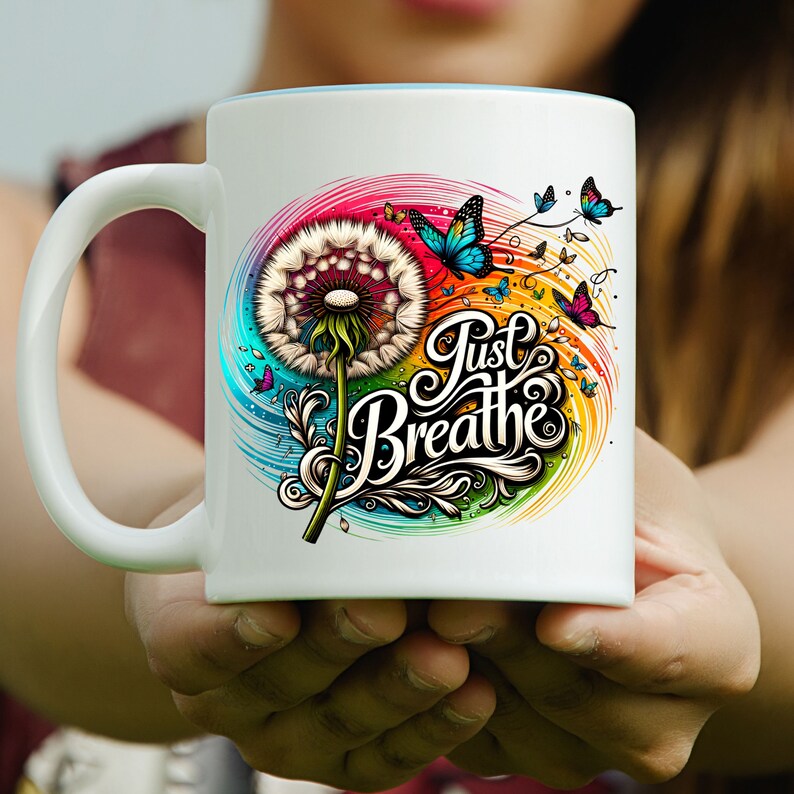 K&ouml;nnte beinhalten: Wei&szlig;e Keramiktasse mit einem bunten Design mit einem L&ouml;wenzahn, Schmetterlingen und dem Text "Just Breathe".