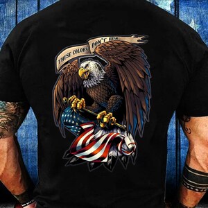 May include: Black t-shirt featuring a detailed illustration of an American bald eagle clutching a US flag. The eagle is brown, white, and yellow, with the text "These Colors Don't Run" above it. The flag has red and white stripes and blue stars.