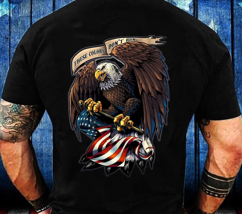 May include: Black t-shirt featuring a detailed illustration of an American bald eagle clutching a US flag. The eagle is brown, white, and yellow, with the text "These Colors Don't Run" above it. The flag has red and white stripes and blue stars.