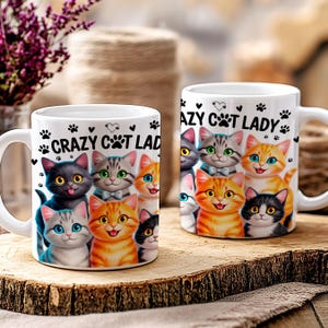 May include: Two white ceramic mugs with the text "Crazy Cat Lady" and a colorful illustration of various cat breeds. The mugs feature a handle and are decorated with paw prints and hearts. The mugs are displayed on a wooden surface.