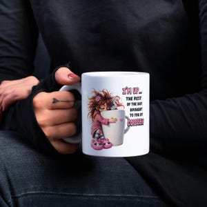 K&ouml;nnte beinhalten: Wei&szlig;e Keramik-Kaffeetasse mit einer Cartoon-Illustration eines M&auml;dchens, das eine Kaffeetasse umarmt. Auf der Tasse steht der Text "I'M UP... THE REST OF THE DAY BROUGHT TO YOU BY COFFEE!" in Rosa und Wei&szlig;.
