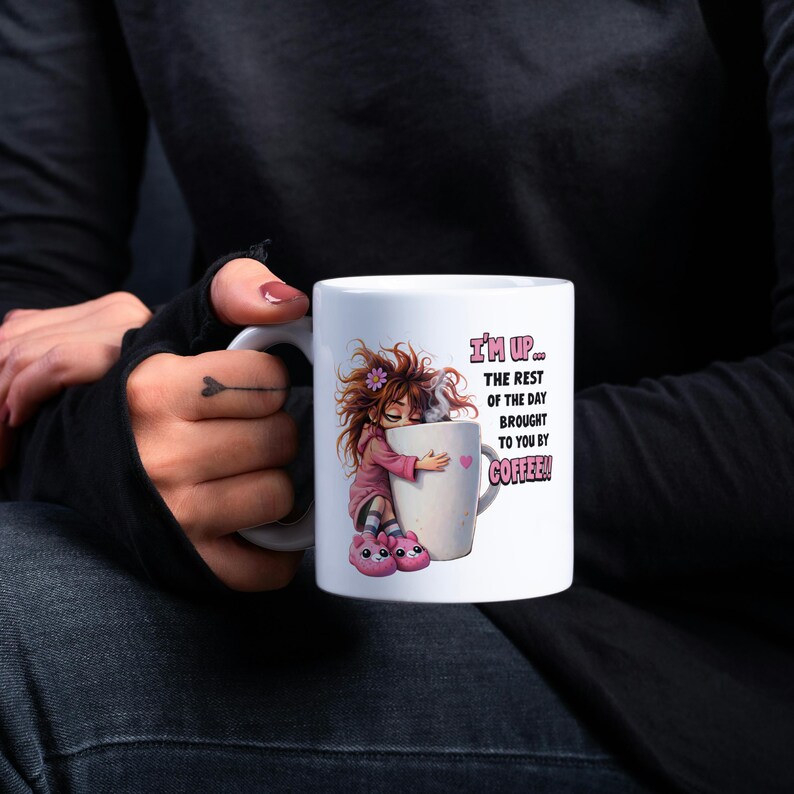 K&ouml;nnte beinhalten: Wei&szlig;e Keramik-Kaffeetasse mit einer Cartoon-Illustration eines M&auml;dchens, das eine Kaffeetasse umarmt. Auf der Tasse steht der Text "I'M UP... THE REST OF THE DAY BROUGHT TO YOU BY COFFEE!" in Rosa und Wei&szlig;.