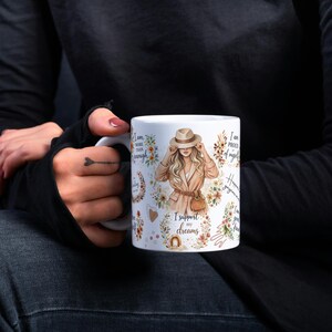 May include: White ceramic mug featuring a woman in a tan coat and hat, surrounded by floral designs and text. The mug includes the phrases "I am more than enough" and "I support my dreams."
