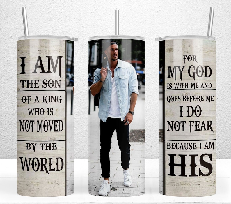 May include: A stainless steel tumbler with a straw, featuring a man in a light blue shirt and black pants. The tumbler has text that reads "I AM THE SON OF A KING WHO IS NOT MOVED BY THE WORLD" and "FOR MY GOD IS WITH ME AND GOES BEFORE ME I DO NOT FEAR BECAUSE I AM HIS."