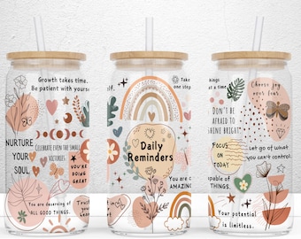 Op de afbeelding: Drie glazen bekers met bamboe deksels en rietjes. Elke beker heeft een uniek ontwerp met inspirerende citaten, regenbogen, vlinders en bloemelementen in tinten perzik, beige en bruin. De tekst bevat zinnen als "Daily Reminders".