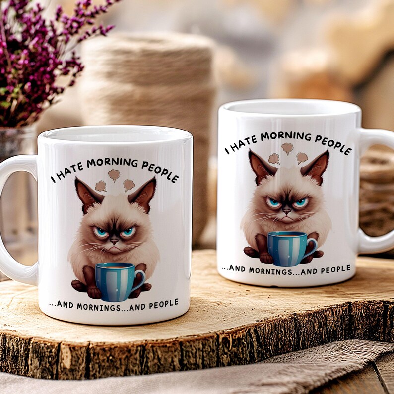 Puede incluir: Dos tazas de cer&aacute;mica blanca con una ilustraci&oacute;n de un gato gru&ntilde;&oacute;n y el texto "I HATE MORNING PEOPLE...AND MORNINGS...AND PEOPLE". El gato tiene una taza azul.
