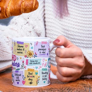 May include: White ceramic mug with colorful text and illustrations. The mug features phrases like "Shame makes it worse" and "Task paralysis is real." Floral and heart designs decorate the mug. A croissant is held above the mug.