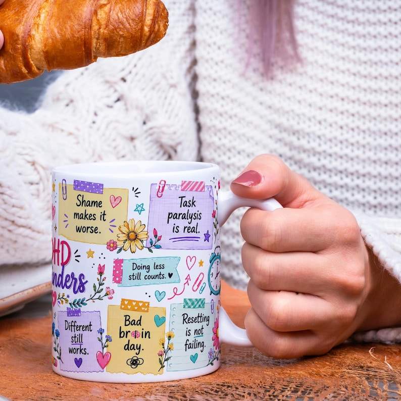 May include: White ceramic mug with colorful text and illustrations. The mug features phrases like "Shame makes it worse" and "Task paralysis is real." Floral and heart designs decorate the mug. A croissant is held above the mug.