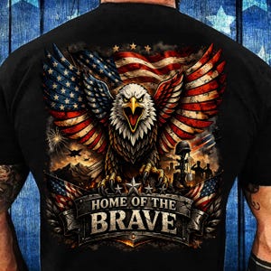 May include: Black t-shirt featuring a detailed graphic of an American bald eagle with outstretched wings, incorporating the American flag's stars and stripes. The design includes the text "HOME OF THE BRAVE" on a banner, with military imagery.