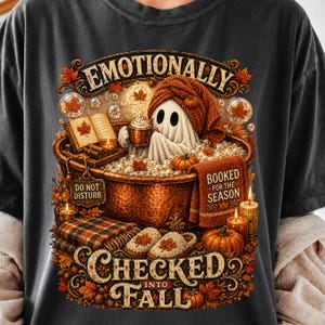 May include: A charcoal grey t-shirt featuring a graphic of a ghost in a copper bathtub filled with bubbles. The ghost wears a knit cap and holds a mug. Text reads EMOTIONALLY CHECKED INTO FALL and BOOKED FOR THE SEASON. Includes pumpkins and leaves.
