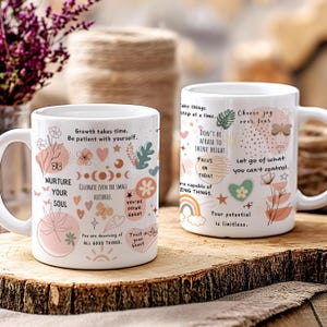 K&ouml;nnte beinhalten: Zwei wei&szlig;e Keramikbecher mit inspirierenden Zitaten und floralen Designs. Ein Becher zeigt Texte wie "Nurture Your Soul" und "Celebrate Even the Small Victories". Der andere Becher hat S&auml;tze wie "Choose Joy Over Fear" und "Your Potential is Limitless".