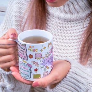 Pode incluir: Caneca de cer&acirc;mica branca com ilustra&ccedil;&otilde;es coloridas e texto. A caneca apresenta um arco-&iacute;ris, flores e a palavra "Different". O texto inclui "Remember teaching is a work of" e "Reminder: the influence of a good teacher cannot be erased."
