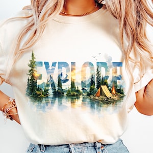 May include: Cream-colored t-shirt featuring the word "EXPLORE" in blue, with a scenic landscape design. The design includes mountains, trees, a lake, and a tent with a campfire, evoking a sense of adventure and the outdoors.