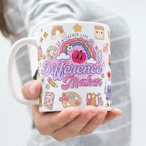 Puede incluir: Taza de cerámica blanca con un diseño envolvente con el texto "Difference Maker" en una fuente retro. El diseño incluye un arcoíris, una manzana, un lápiz y otras ilustraciones con temática escolar. La taza está sostenida por una persona.