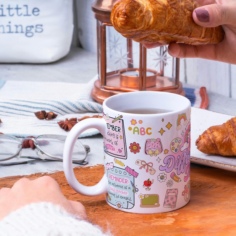 Pode incluir: Caneca de cer&acirc;mica branca com um design colorido com a palavra "Different" e ilustra&ccedil;&otilde;es com tema escolar. A caneca cont&eacute;m uma bebida quente. Um croissant &eacute; segurado perto.