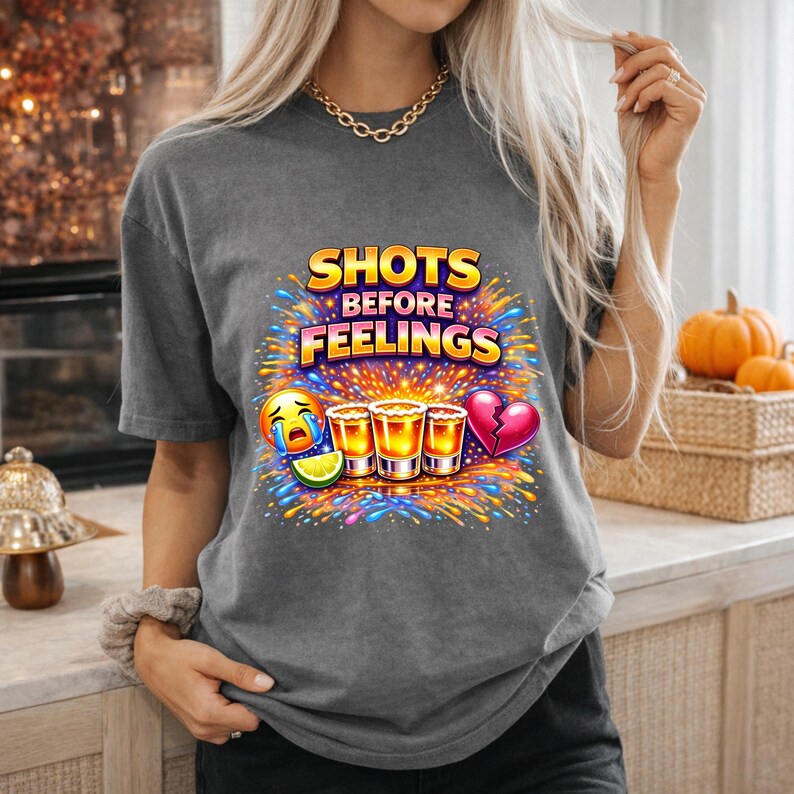 Pu&ograve; includere: T-shirt grigio con la scritta "SHOTS BEFORE FEELINGS" in giallo e blu. La grafica include bicchierini, un'emoji che piange, una fetta di lime e un cuore spezzato. Il design &egrave; circondato da fuochi d'artificio colorati.