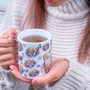 K&ouml;nnte beinhalten: Wei&szlig;e Keramiktasse mit floralem Design mit Schmetterlingen und inspirierenden Zitaten. Die Tasse ist mit einer hellbraunen Fl&uuml;ssigkeit gef&uuml;llt und wird von einer Person gehalten, die einen wei&szlig;en Pullover tr&auml;gt.