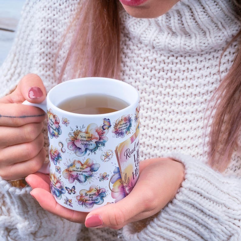 K&ouml;nnte beinhalten: Wei&szlig;e Keramiktasse mit floralem Design mit Schmetterlingen und inspirierenden Zitaten. Die Tasse ist mit einer hellbraunen Fl&uuml;ssigkeit gef&uuml;llt und wird von einer Person gehalten, die einen wei&szlig;en Pullover tr&auml;gt.