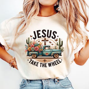 May include: A white t-shirt with a graphic of a vintage pickup truck with flowers and cacti. The text "JESUS TAKE THE WHEEL" is printed below the truck.