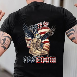 May include: Black t-shirt featuring a patriotic design. The graphic includes an American flag, an eagle with outstretched wings, and military boots. The words "ROOTS OF FREEDOM" are displayed in a bold font.