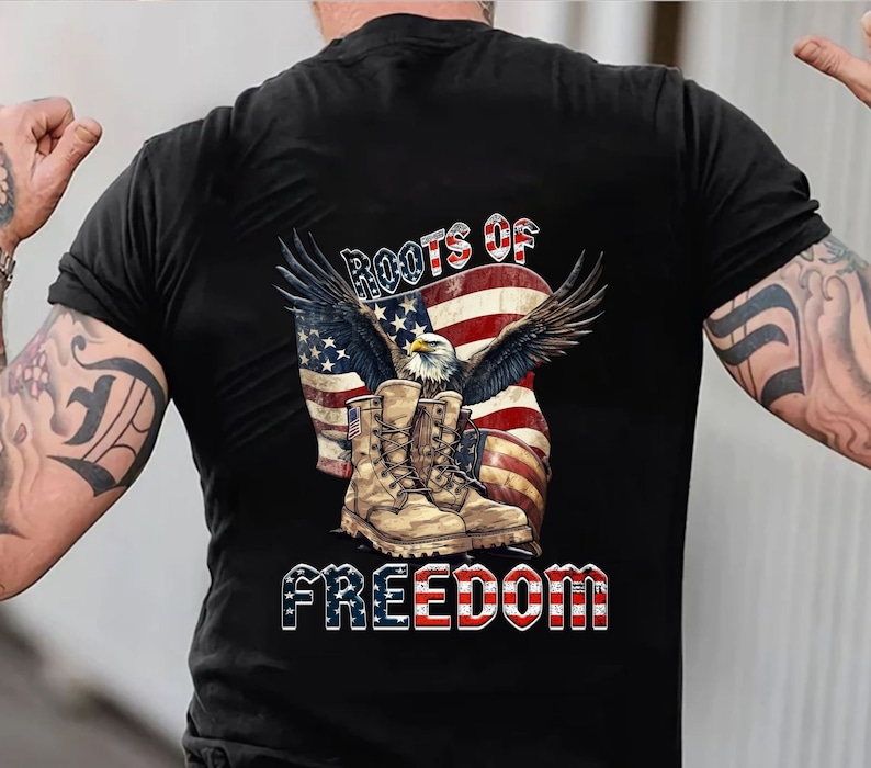May include: Black t-shirt featuring a patriotic design. The graphic includes an American flag, an eagle with outstretched wings, and military boots. The words "ROOTS OF FREEDOM" are displayed in a bold font.
