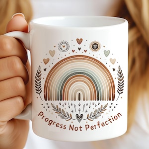 Könnte beinhalten: Weiße Keramiktasse mit einem Regenbogen-Design in Braun-, Beige- und Grüntönen. Der Regenbogen ist von kleinen Herzen und Blättern umgeben. Der Text "Progress Not Perfection" ist auf die Tasse gedruckt.