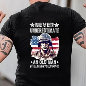May include: Black t-shirt featuring a portrait of a soldier in a helmet, superimposed over a distressed American flag. The text reads "NEVER UNDERESTIMATE AN OLD MAN WITH A MILITARY BACKGROUND" in white.