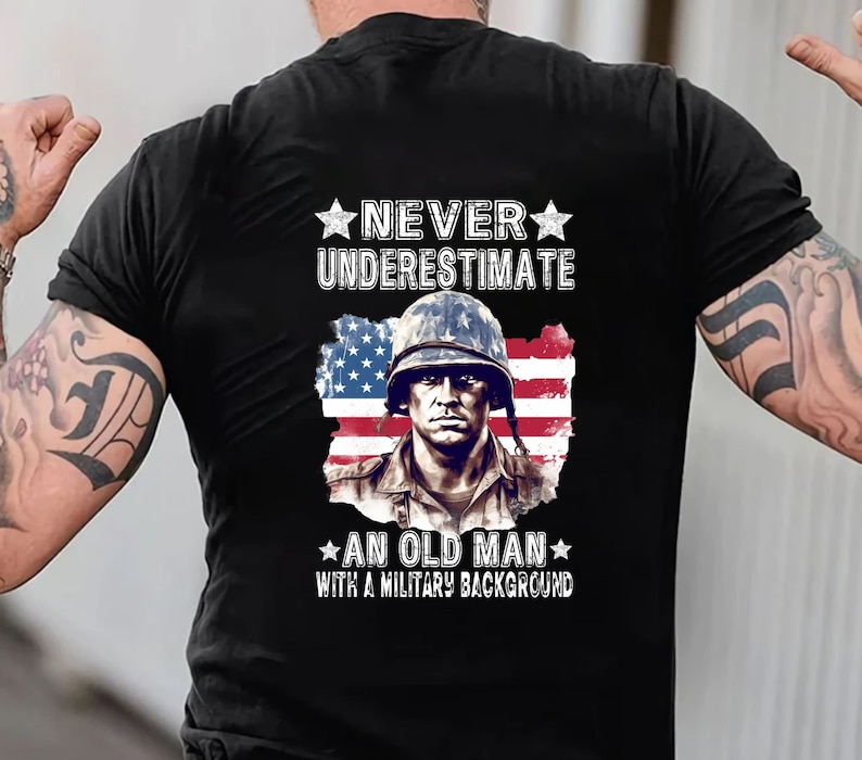 May include: Black t-shirt featuring a portrait of a soldier in a helmet, superimposed over a distressed American flag. The text reads "NEVER UNDERESTIMATE AN OLD MAN WITH A MILITARY BACKGROUND" in white.