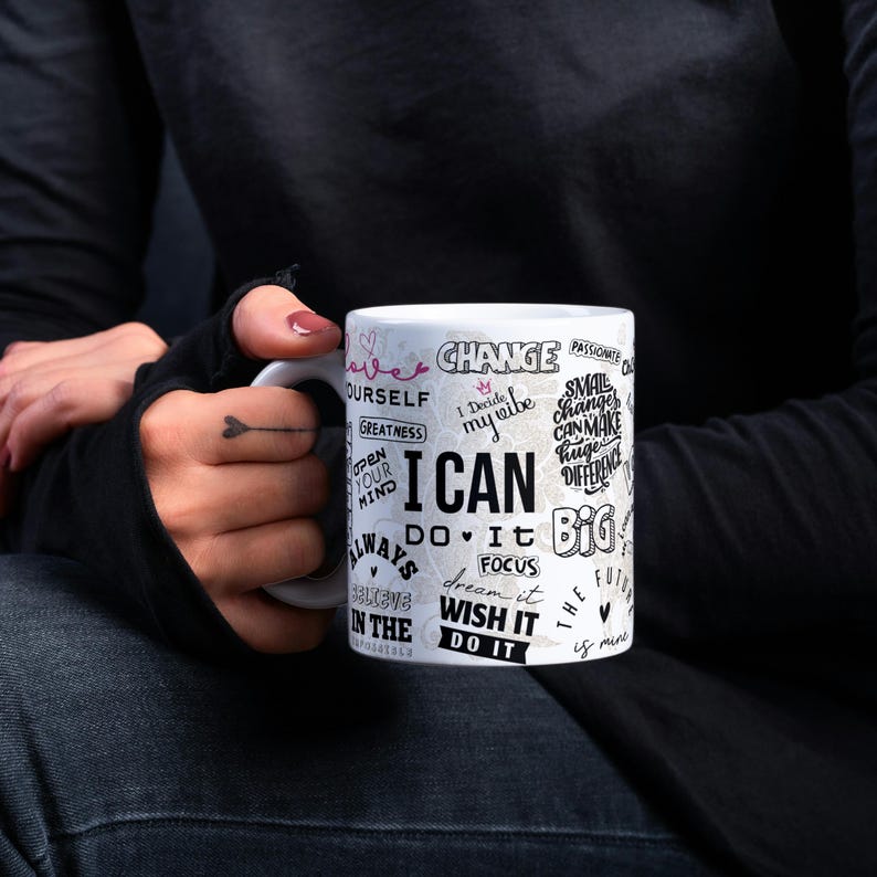 May include: White ceramic mug with the words "I CAN" in large black letters. The mug is covered in motivational phrases and words like "change", "dream it", and "focus". The mug is held by a person wearing a black long-sleeve shirt.