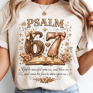 May include: Beige t-shirt featuring the text "PSALM 67" with decorative floral and bow accents. The design includes an open book and the phrase "God be merciful unto us, and bless us; and cause his face to shine upon us,"