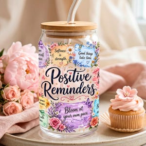 Peut inclure: Un gobelet en verre transparent avec un couvercle en bois et une paille. Le gobelet est décoré de motifs floraux et des mots "Positive Reminders". Le design comprend les phrases "Bloom at your own pace" et "Good things take time."