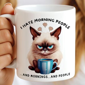 Op de afbeelding: Witte keramische mok met een chagrijnige cartoonkat met blauwe ogen, die een blauwe koffiekop vasthoudt. De mok heeft de tekst "I HATE MORNING PEOPLE...AND MORNINGS...AND PEOPLE" in het zwart.
