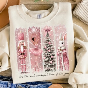 May include: Cream-colored sweatshirt featuring a festive design with a pink nutcracker, ballerina, Christmas tree, and rabbit. The design includes the text "it's the most wonderful time of the year". The graphic has a winter theme.