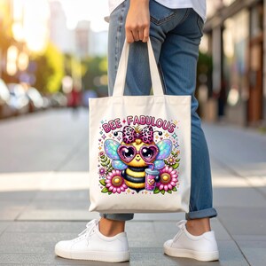 May include: A cream-colored tote bag featuring a colorful cartoon bee with heart-shaped sunglasses, a bow, and the words "BEE FABULOUS." The bee is surrounded by flowers and holding a drink. The bag is being held by a person wearing jeans and white sneakers.