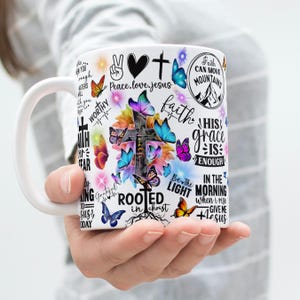 May include: White ceramic mug with a cross, butterflies, and floral designs. The mug is decorated with inspirational phrases such as "Faith Can Move Mountains" and "His Grace Is Enough". The design incorporates bright colours and various fonts.