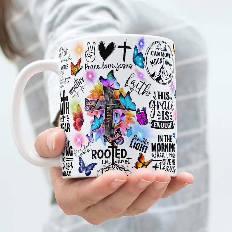 May include: White ceramic mug with a cross, butterflies, and floral designs. The mug is decorated with inspirational phrases such as "Faith Can Move Mountains" and "His Grace Is Enough". The design incorporates bright colours and various fonts.