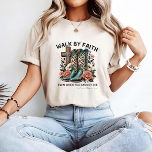May include: A beige t-shirt with a graphic of cowboy boots, a cross, and flowers. The text "Walk by Faith" is above the graphic and the text "Even when you cannot see 2 Corinthians 5:7" is below the graphic.