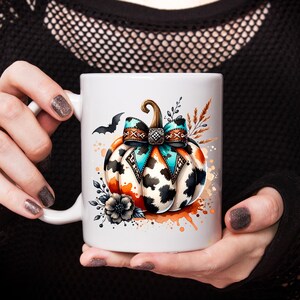 May include: White ceramic mug featuring a watercolor design of a pumpkin with a turquoise and brown bow, black and white cow print, and a bat. The pumpkin has orange accents and is surrounded by floral elements.