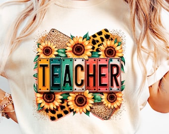 May include: Cream-colored t-shirt featuring the word "TEACHER" in colorful license plate-style letters, surrounded by sunflowers and leopard print accents. The design is a vibrant mix of colors, perfect for a teacher.