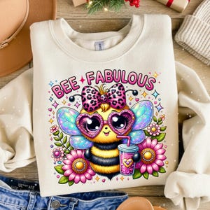 May include: Cream-colored sweatshirt with a colorful graphic of a bee wearing heart-shaped glasses and a bow. The bee holds a drink and is surrounded by flowers. The text "BEE FABULOUS" is above the bee in pink letters.