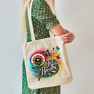 K&ouml;nnte beinhalten: Eine wei&szlig;e Canvas-Tragetasche mit einem bunten Design aus L&ouml;wenzahn und Schmetterlingen. Der Text "Just Breathe" ist in einer stilisierten Schriftart geschrieben.
