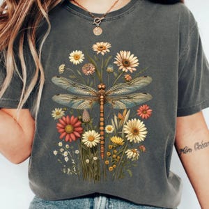 May include: A charcoal gray short-sleeve t-shirt featuring a central graphic of a large, detailed dragonfly with iridescent wings. The dragonfly is surrounded by a vibrant arrangement of various wildflowers in shades of red, yellow, and white.