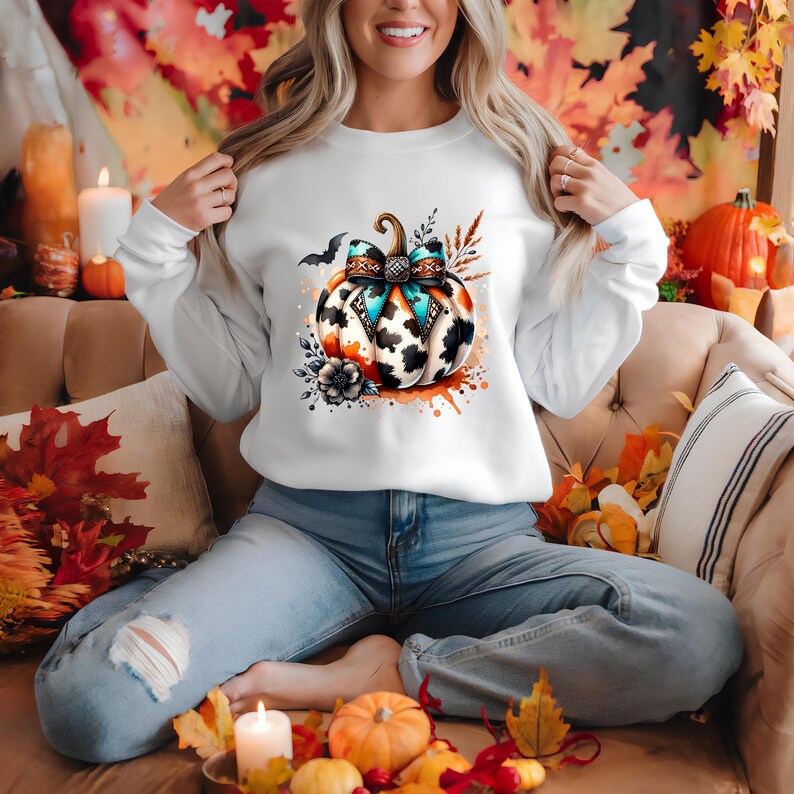 May include: White long-sleeve sweatshirt featuring a watercolor pumpkin design. The pumpkin has a black and white cow print, a turquoise and black bow, and orange accents. A black bat and floral elements complete the design. The sweatshirt is a fall-themed item.